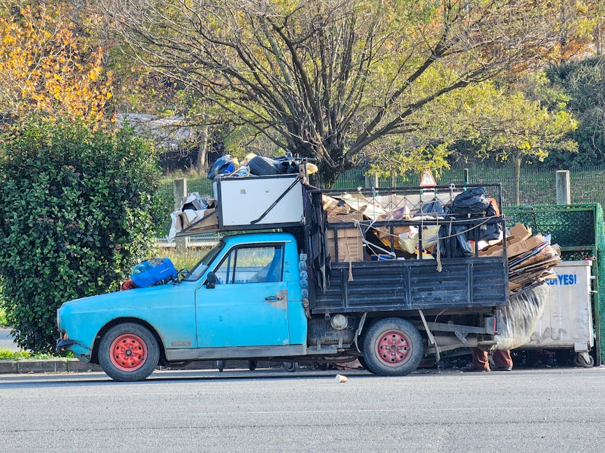 A light blue, small flatbed truck parked along a suburban street, heavily loaded with various types of rubbish and discarded materials. The cargo includes large black plastic bags, flattened cardboard