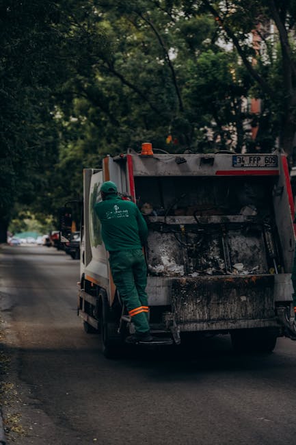 A worker dressed in a green uniform and safety helmet is standing beside the open rear of a commercial rubbish collection van, which is parked on a residential street lined with trees. The van, showin
