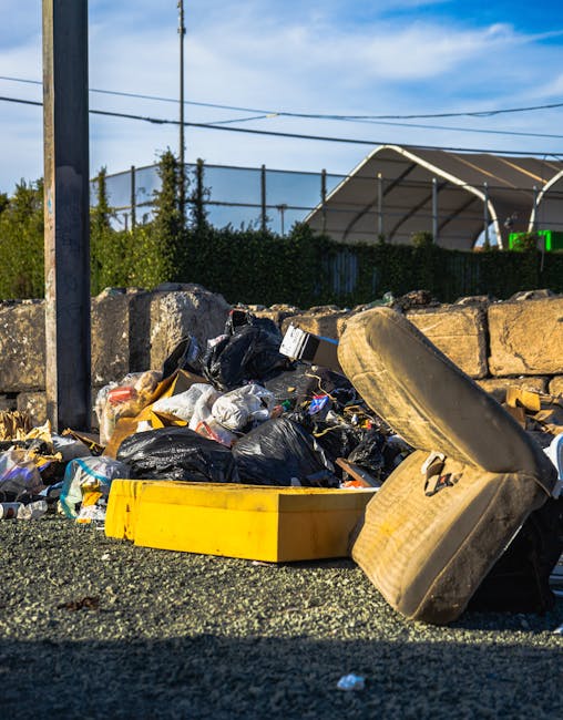 A collection of overflowing rubbish bins and scattered waste on a paved urban street scene in front of a commercial building with blue scaffolding. The waste includes black, red, and grey wheelie bins